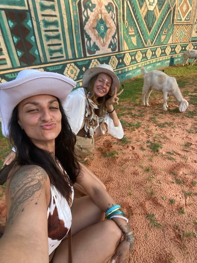 Two women are photographed in Mui Ne, Vietnam, in front of a colorful, tapestry-like mural depicting geometric patterns and stylized figures. The woman in the foreground, with dark hair and wearing a white cowboy hat and a cow-print bikini top, is looking directly at the camera and making a duck-face. She has extensive tattoos on her left arm and shoulder. The woman in the background, also wearing a white cowboy hat, has blonde hair and is smiling at the camera, holding up two fingers in a peace sign. She is wearing a white top with feather-like embellishments around the neckline and a tan crossbody bag. Behind them, two goats are visible on a patch of grass. The ground in the foreground appears to be reddish sand with sparse green vegetation. The overall setting suggests an outdoor, possibly festive or tourist-oriented location in Mui Ne.