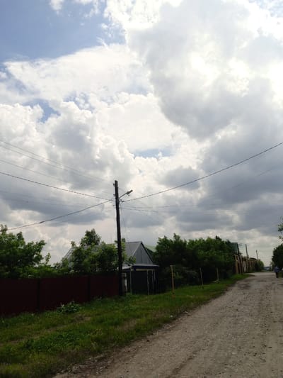 This is a daytime, outdoor, eye-level, medium shot of a dirt road in Pavlovsk, Russia, on a cloudy day. The road is on the right side of the image and appears to be unpaved, with gravel and dirt. On the left side of the road, there is a reddish-brown metal fence, followed by a patch of green grass and vegetation. Behind the fence, there are trees and the roofs of houses. A wooden utility pole with electrical wires runs horizontally across the upper portion of the image. The sky is filled with large, white, and gray clouds. A lone person in blue clothing is walking away from the camera on the road in the distance on the right.