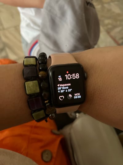 The image shows a close-up of a person's wrist wearing a smartwatch and two beaded bracelets. The smartwatch displays the time as 10:58 and shows weather information for Ikryanoye, Russia, indicating a temperature of 29 degrees Celsius and clear skies, with a projected high of 33 degrees and a low of 22 degrees. Below the weather information, a heart icon and a circular icon representing signal strength are visible. The smartwatch also displays "MCK" and "09:58", possibly indicating a local time or another metric. The bracelets are made of various colored beads, including purple, green, and dark brown, with one featuring a tiger's eye stone. The background is blurred, but appears to be an indoor setting with hints of clothing and furniture. The person's skin is visible and appears to have hair.