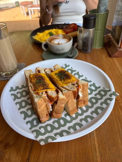 A top-down, slightly angled shot depicts a breakfast or brunch scene on a wooden table. The primary focus is a white plate lined with patterned green and white paper, holding a cut sandwich. The sandwich features toasted bread, a filling of shredded vegetables, a protein (possibly fried chicken or fish), and a slice of melted cheese, possibly cheddar, with a burnt or charred appearance. To the left of the plate, there is a cup of coffee with latte art, next to a small plate of scrambled eggs. A salt and pepper shaker set is also present. Part of a person's arm and clothing is visible on the left side of the frame. A glass containing a beverage (likely a latte or iced coffee) sits in the lower left corner. The location is Pecatu, Indonesia.