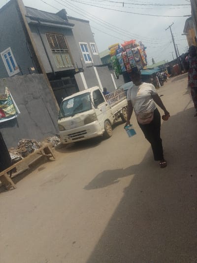 On a sunlit street in Agani, Nigeria, an adult female is observed walking towards the viewer, slightly off-center to the right. She wears a light-colored short-sleeved top, dark trousers, and sandals, with a pink fanny pack positioned at her waist. A large, multi-tiered display of assorted packaged snacks, primarily yellow and blue, is balanced on her head. In her right hand, she carries an orange-colored fruit, and in her left, a light blue plastic container. Her shadow projects on the asphalt road to her left.
To the left of the frame, a white, compact utility pickup truck, likely a Daihatsu Hijet or similar model, is parked facing away from the viewer. The truck shows minor damage to its front bumper and grille. Behind the truck stands a two-story building painted dark gray, featuring white-framed windows and a second-floor balcony secured by gold-colored security bars. A concrete wall partially visible on the far left displays a large political poster. Adjacent to this wall, a wooden bench or stand is piled with miscellaneous debris and discarded items. In the background, other structures, power lines, and additional individuals are discernible, suggesting a commercial or market district. The sky is partially overcast.