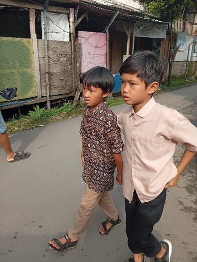 Two young boys walk down a narrow street. The boy on the left wears a patterned brown shirt, tan pants, and sandals. The boy on the right wears a light pink shirt, black pants, and sandals. They both look to their left. Dilapidated buildings line one side of the street with patches of green growth. Another person in sandals walks in the periphery of the image.