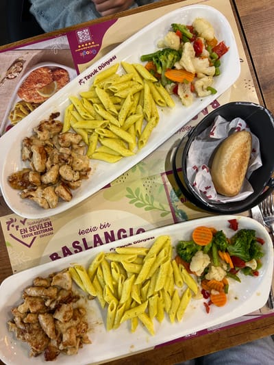 The image shows two white oval plates of food, likely from a restaurant, on a wooden table. The top plate contains penne pasta seasoned with herbs and a side of mixed steamed vegetables including broccoli, cauliflower, carrots, and bell peppers. The bottom plate also contains penne pasta and a generous portion of grilled chicken pieces. To the right of the bottom plate, a small black metal bucket lined with paper holds a single bread roll. The background includes placemats with Turkish text and branding for "Tavuk Dünyası," which translates to "Chicken World," a popular Turkish restaurant chain. The overall scene suggests a casual dining experience. The presence of specific restaurant branding, "Tavuk Dünyası," and the mention of "Şişli, Turkey" in the prompt, implies the location of this meal.