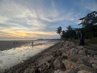 The image shows a beach scene in Baan Sri Thanu, Thailand, during sunset. The tide is low, revealing a wide expanse of wet sand reflecting the colors of the sunset. Several people are walking on the beach. Along the shoreline, there is a barrier made of large rocks. A person wearing a long dress stands near some trees and vegetation. Palm trees are visible in the background. The sky is filled with clouds tinged with yellow and orange hues from the setting sun.