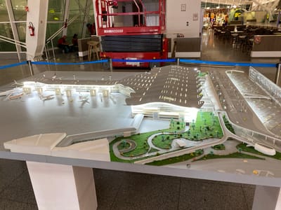 The image shows a detailed architectural model of an airport in a modern, indoor setting. The model features a long, curved terminal building with a distinctive roof design, complete with rows of small, illuminated windows. Surrounding the terminal are various elements of an airport, including a tarmac with several model airplanes, access roads, parking lots, and a landscaped area with miniature trees and pathways. In the foreground, the model is displayed on a white pedestal.
In the background, the actual airport terminal is visible. There are large windows, a red scissor lift labeled "MANIACCESS 120 SE" with two individuals working on it. The floor appears to be polished concrete. To the right of the scissor lift, there are tables and chairs set up in a seating area, with shops or concessions visible in the distance. A blue cord barrier separates the model from the operational area.
The scene is consistent with a display or exhibit within the Houari Boumediene Airport in Dar El Beïda, Algeria, given the scale and style of the architectural model and the typical airport environment depicted.