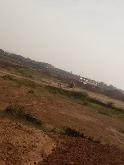 The image depicts a dusty, open terrain with scattered vegetation and some construction activity. The ground is predominantly red-brown soil, indicative of arid or semi-arid conditions. In the mid-ground, there is an excavator, a heavy machinery used in construction, accompanied by a group of people. Further in the background, there are several structures, likely houses or buildings, suggesting a developing settlement or a rural area. The overall environment appears to be an outdoor scene under a hazy sky. The tilt of the camera suggests the image was taken on a slope or at an angle. This scene is located in Kwang, Nigeria.
