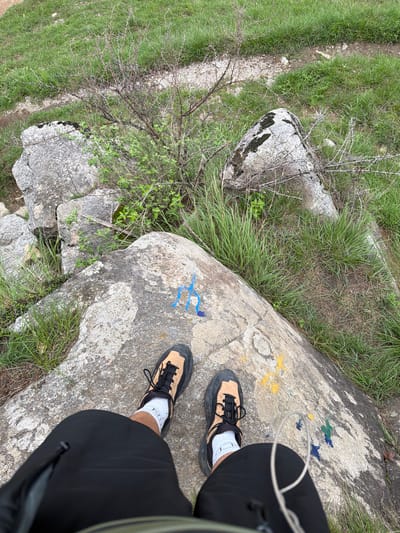 The image depicts a first-person perspective of a person standing on a large, weathered rock, likely in a natural outdoor setting. The rock has patches of moss and lichen on its surface and is marked with several colorful spray-painted symbols, including blue and yellow. The person is wearing black athletic shorts, white socks, and tan and black hiking shoes. In the background, there is green grass, dry brush, and more rocks, suggesting a rugged or mountainous terrain. The overall scene conveys a sense of exploration or hiking. The location is identified as Bahang, India.