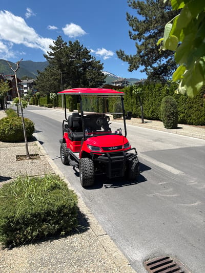 A red "Moleto" golf cart is parked on a paved road in Razlog, Bulgaria. The road is flanked by trimmed hedges and trees, with mountains visible in the background. The sky is blue with scattered clouds. A storm drain is visible on the edge of the road.
