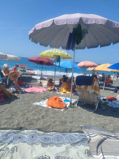 This is a daytime, eye-level, outdoor photograph taken on a beach in Lavagna, Italy. The scene depicts a sunny day with a clear blue sky and calm ocean in the background. Several beach umbrellas are set up on a pebbly shore, providing shade for beachgoers. In the foreground, a woman with gray hair is sitting in a blue beach chair, holding a straw hat in her lap. She is wearing a black swimsuit. To her left, the legs of another person are visible, suggesting they are also seated. Various beach towels and bags are scattered on the sand.

Further back, a group of people are enjoying the beach. Some are lying on towels, while others are sitting or standing near the water's edge. Children appear to be present in the water, where small waves are breaking. The ocean is a vibrant blue, and a few sailboats are visible in the distance. The overall atmosphere is one of relaxation and recreation on a typical summer day at the Italian coast.