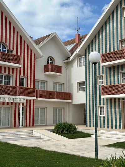 This image depicts an exterior view of a residential or commercial complex in Ílhavo, Portugal, characterized by its distinctive striped architectural cladding.
The scene is dominated by three interconnected building sections, all featuring pitched roofs with reddish-brown tiles and white trim along the eaves. The leftmost building section is clad in vertical red and white stripes. It displays multiple rectangular windows with white frames and two balconies with reddish-brown latticework railings. The central building section is predominantly white/cream-colored, featuring various rectangular windows and one prominent arched window with a half-circle top. This section also includes at least two balconies with matching reddish-brown latticework railings. A chimney with an antenna extends from the roofline above this central area. The rightmost building section is clad in vertical teal/green and beige/white stripes. It shows multiple rectangular windows with white frames and two balconies with reddish-brown latticework railings.
The ground level features a paved courtyard or walkway system. Sections of manicured green grass are visible in the foreground and bordering the paved areas. A small, circular cluster of green shrubbery is centrally located within the paved space. To the left, white-colored steps lead up, adjacent to a small ramp. A dark green street lamp with a spherical white globe fixture stands vertically in the midground on the right side of the paved area.
The sky overhead is partly cloudy, with areas of clear blue interspersed with white clouds, indicating daylight conditions. No individuals are visible, and no specific actions or interactions are depicted.