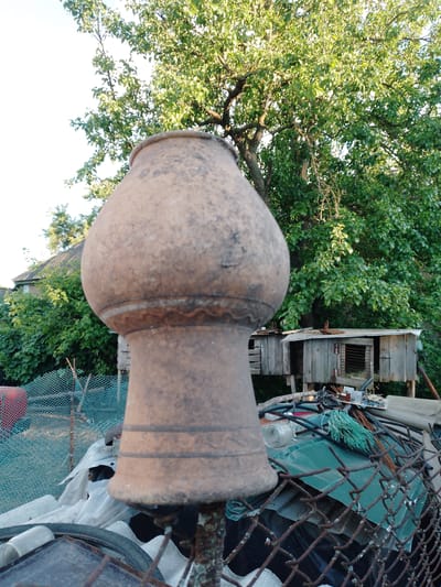 The image captures a rustic outdoor scene, dominated in the foreground by a large, out-of-focus, earthy brown ceramic pot or finial. Its textured surface shows signs of age and weathering, featuring horizontal ridges and a bulbous shape.
The background reveals a vibrant green canopy of leaves from a large tree, possibly a fruit tree, with sunlight filtering through, creating dappled light and shadow. To the left, a portion of a house or shed with a grey, possibly corrugated, roof is visible amidst dense foliage. Further right, a weathered wooden structure, resembling an animal hutch or small enclosure, stands on stilts.
Below the main subject and extending into the midground, a turquoise mesh fence runs horizontally, partially obscured by various items. Accumulated beneath and around the fence are miscellaneous objects, including corrugated metal sheets (some green, some rusty), tangled wires, a circuit board, plastic containers, a bicycle tire, and white plastic sheeting, suggesting a storage or utility area in a backyard.
The scene appears to be set during daytime, likely late afternoon or early morning, given the soft, natural lighting and lack of harsh shadows. The weather is clear and sunny. No people are visible, and no specific activities are taking place; the image presents a static view of a backyard. There is no visible text. The overall setting evokes a semi-rural or peri-urban environment, consistent with a backyard in an area like San Martín de Porres, Peru.