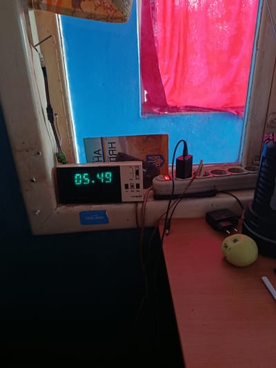 The image shows a close-up view of a window sill with various objects on it. A digital clock displays the time as 05:49. To the right of the clock is a power strip with a red indicator light on and two black power adapters plugged in. A small green apple is positioned on the right side of the frame. In the background, a bright pink curtain is visible through the window, along with a blue painted wall. A book with Cyrillic text is partially visible behind the clock. The overall scene appears to be an indoor setting, possibly a residential room, with a focus on electrical devices and everyday items. The image does not contain any identifiable landmarks or contextual clues that would place it specifically in Kremenchuk, Ukraine.