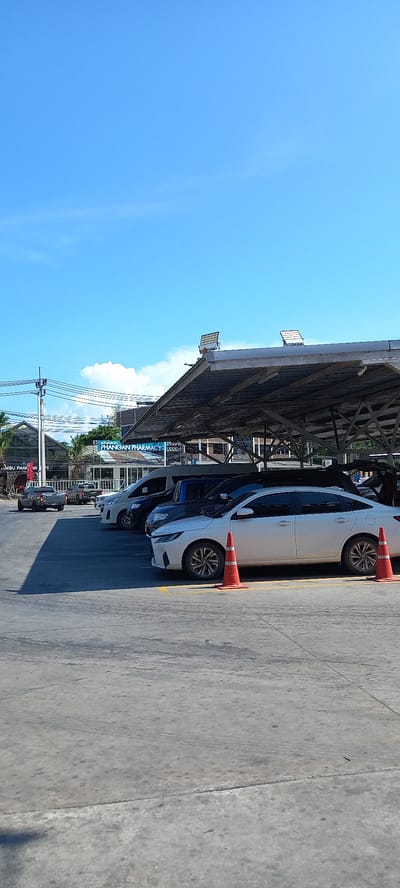 This image captures a bright, sunny daytime scene in what appears to be a commercial area or parking lot in Moo 1, Thailand. The sky is a clear, vibrant blue with a few scattered white clouds.

In the foreground, a concrete or asphalt surface serves as a parking area, marked with yellow lines. Several cars are parked parallel to the viewer, predominantly sedans; a white car with distinctive black and silver alloy wheels is most prominent, accompanied by at least one darker-colored vehicle. Orange traffic cones are placed next to the wheels of the white car.

An overhead structure, resembling a large metal-framed canopy or the underside of a building's extension, provides shade over the parked vehicles. Utility poles and power lines crisscross the mid-ground, indicating a developed streetscape.

In the background, beyond the parked cars and utility infrastructure, other buildings and tropical greenery are visible. A clear blue sign with white lettering is prominent, reading "PHANGAN PHARMACY," strongly suggesting the location is Koh Phangan, Thailand. Below this, another partially visible sign appears to begin with "KO SAMUI PHAR," possibly indicating another branch or related business.

The scene depicts a calm, ordinary moment with no people visible or specific events taking place. The weather is clear and bright, characteristic of a sunny day.