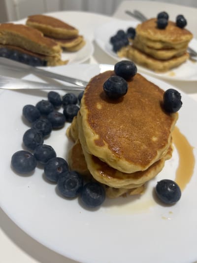 The image shows three white plates of pancakes garnished with blueberries. Each plate has a stack of pancakes and a scattering of blueberries. Syrup is drizzled on one plate. Silverware is visible near the pancakes. The setting is a bright, indoor location. There is no indication of Mogán, Spain in this image.
