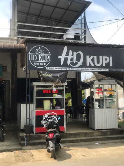 The image depicts a street view of a small food stall and cafe named "HD KUPI" in Muliengmanyang, Indonesia. The cafe has a prominent black sign with white lettering displaying the name "HD KUPI" and "Free Wi-Fi". Below this, an address is visible: "Jl. Buloh Beureughang, Gp. Cot Seutui, Kec. Kuta Makmur - Aceh Utara". A phone number, "HP: 0822 7607 7302", is also displayed on the sign.

The food stall is set up in front of the cafe, featuring a glass display case with various food items and menus. Red and black signage indicates offerings such as "AYAM PENYET" (smashed chicken), "NASI GORENG" (fried rice), and "MIE AYAM" (chicken noodles). A person, likely an employee, is visible behind the counter of the food stall. In front of the stall, a black motorcycle is parked.

The environment appears to be a typical Indonesian street scene with concrete pathways and a bright, sunny sky overhead. The architecture of the cafe suggests a simple, open-air structure with a metal roof. The overall impression is of a casual, local establishment catering to passersby.