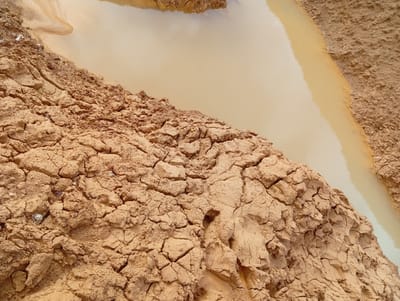 The image shows a close-up, overhead view of a muddy water puddle next to cracked, dry earth. The water is a cloudy, light brown color, reflecting the sky and surrounding earth. The dry earth is a reddish-brown hue and exhibits a network of deep cracks, indicating a dry, arid environment. The texture of the earth is rough and uneven, with some small stones and debris visible. The scene is likely located in Kuru, Nigeria, where such geological conditions can be found. There are no people or animals present in the image.