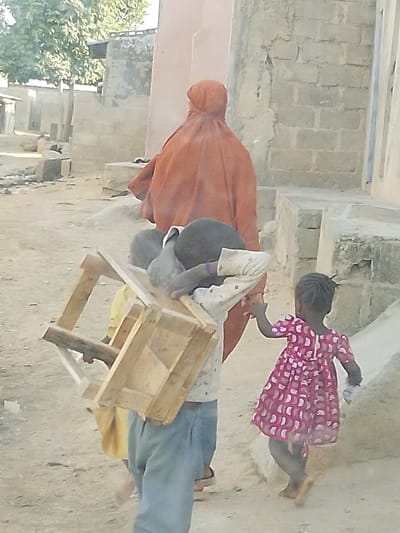 A woman wearing an orange burqa walks away, followed by three children. Two kids carry a wooden structure, possibly a chair or stool, on their shoulders. A small girl in a pink patterned dress holds the hand of one of the boys carrying the wooden object. They are on a dusty path between buildings constructed of concrete blocks. A tree is visible in the background.