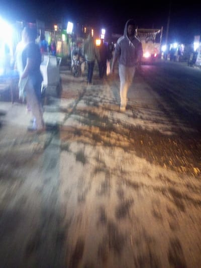 A blurry, low-quality image depicts a nighttime street scene. Several people are visible, including a hooded figure walking towards the camera. Other individuals are near vendor stalls with bright lights. A vehicle, potentially a truck, stands in the background. The ground appears to be cracked or uneven with patches of light and shadow. The overall image suggests a busy, urban environment at night.