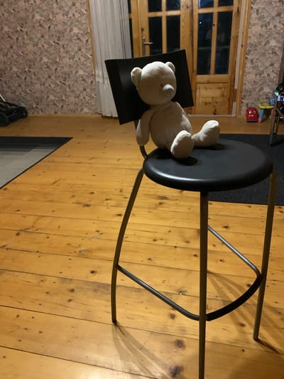 A light beige teddy bear is seated on a black stool in a room with wooden floors. The stool has three metal legs and a round black seat. The teddy bear is facing left and appears to be leaning against a black object that is partially visible behind it. In the background, there is a wooden door with glass panes and a patterned wallpaper. To the left of the door, a white curtain is visible. The floorboards are laid horizontally and appear to be made of light-colored wood. The scene is lit by artificial light, casting shadows on the floor. The location is Mepiskalaki, Georgia.