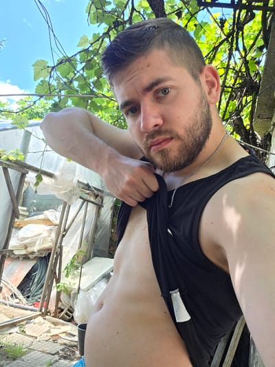 A man with short, dark hair, a beard, and a silver chain around his neck is holding up the bottom of his black tank top to reveal his torso. He appears to be in an outdoor area. Behind him is a structure constructed from white and metal materials, possibly a partial enclosure. Foliage, including grape vines, is visible against a background of blue sky. The location is in a yard near Yagodovo, Bulgaria.