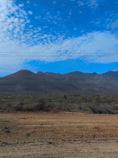 The image shows a landscape in La Cruz del Pastel, Venezuela. The foreground features a barren, brown, dirt terrain. Sparse, dry vegetation is present in the middle ground. A mountain range is in the background under a blue sky with scattered clouds. Overhead power lines stretch across the sky.
