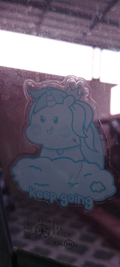 A light blue sticker of a cartoon unicorn wearing a crown sits on a window. The unicorn is depicted as a bust with a smiling face, large eyes, and a horn on its head, all outlined in a darker blue. It is positioned on top of a fluffy cloud, also in light blue. Below the cloud, in the same dark blue outline, are the words "keep going" in a playful font. The sticker is adhered to what appears to be the interior of a vehicle window, indicated by the reflection of the environment outside and the text and symbols common to automotive glass visible at the bottom of the frame. These markings include "TEMPERLITE," "AS2," "2 CIS M," "INO2," and "ADL (211) T," suggesting the vehicle's location in Bandar Lampung, Indonesia, as these are typical markings found on automotive glass in that region. The overall scene is static, with no people or animals present, and the focus is solely on the sticker and its immediate surroundings on the glass.