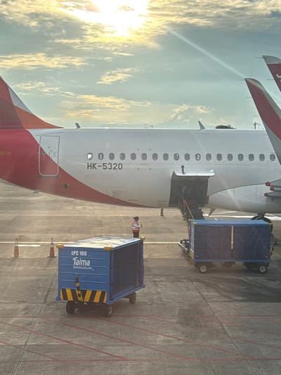 The image showcases an airplane being serviced at an airport. The plane, marked with "HK-5320," has its rear cargo door open. Next to the aircraft are two blue cargo carts, one with diagonal yellow and black stripes. A person wearing a red and white reflective vest stands nearby. The backdrop features a bright, sunlit sky with scattered clouds. The photo is taken from an elevated perspective, possibly from within another aircraft.