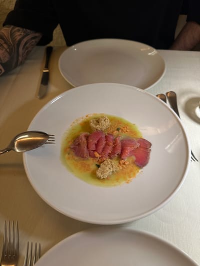 The image shows a white round plate of food on a table setting, likely in a restaurant. The plate contains thinly sliced red meat, possibly tuna or beef carpaccio, arranged on a bed of light-colored sauce, possibly a purée or cream. There are small, irregular pieces of what appears to be croutons or dried bread scattered around the meat. The table is covered with a textured light-colored tablecloth. Adjacent to the plate of food are two other white plates, one on the left and one on the right. A knife is to the left of the main plate, and a fork and spoon are positioned below it. Another set of silverware is visible on the right. A person's arm with a tattoo is visible on the left side of the image. Given the prompt, it can be inferred that this scene is taking place in Agropoli, Italy.