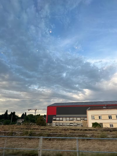 The image depicts an exterior scene, likely in Alès, France, under a partly cloudy sky. In the foreground, a dry, grassy area with some discarded debris is visible, separated by a metal railing. Behind this, a long, multi-story industrial or commercial building is prominent. The building features a dark gray facade with a row of windows across its upper section and a bright red band along its roofline. To the right of this building, a smaller, lighter-colored building with a tiled roof and several windows is situated. Further in the background, beyond a line of trees, a construction crane is visible, suggesting ongoing development or renovation in the area. The sky is a mix of blue and gray clouds, with two bright, circular lights visible in the upper portion of the frame, possibly streetlights or aircraft lights. There are no people or animals visible in the frame.