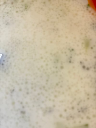 The image is a close-up, blurry shot of a creamy, foamy liquid with small bubbles. There are some darker specks within the foam, suggesting the presence of small solid particles or fragments. In the upper right corner, a sliver of bright orange is visible, possibly a piece of food. The liquid appears to be in a container, suggested by a glint of what might be metal or glass on the far left edge. The background is a pale, off-white color, with a slight greenish tint in some areas, hinting at possible ingredients like herbs or vegetables. The overall impression is of a soup or a blended drink. There is no indication of the city of Vidin, Bulgaria, or any people or specific actions taking place.