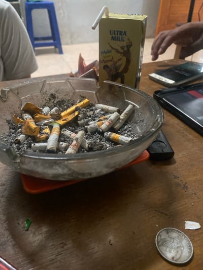 The image captures an indoor scene in the city of Cimenyan, Indonesia, focusing on a cluttered wooden table. A clear, rectangular glass ashtray, heavily laden with dark grey ash, numerous white-filtered cigarette butts, and crumpled gold foil, occupies the center foreground. This ashtray rests on an orange, possibly plastic, base. To its immediate right stands a yellow Ultra Milk carton, featuring the text "Make Your Move!" and an illustration of two figures, with a white, bent drinking straw inserted.
Further right on the table, three mobile devices are present: a black tablet, a black smartphone, and a white smartphone. A black remote control lies beside the ashtray on the right. In the lower right corner of the table, a tarnished silver-colored coin and a small, torn piece of white paper are visible. A small crumpled piece of green material is located in the lower left corner.
The upper left portion of the frame shows the arm and torso of an individual wearing a white shirt, seated at the table. On the right side, the right hand of another individual is visible, positioned near the mobile devices. The background features a light-colored wall, with a blue plastic stool visible in the upper left and part of a wooden door or furniture item in the upper right. The floor, partially visible, appears to be tiled.