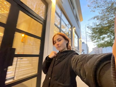 A young adult female, with reddish-brown hair pulled back, is depicted in a self-portrait (selfie) taken at arm's length. She is wearing a black puffer jacket and looks directly at the camera. The scene is an urban exterior, specifically outside "NI. Restaurant" located at 5 Rue Saint-Symphorien in Reims, France.
To the subject's left, a building facade with large, black-framed glass windows is visible. Warm yellow-orange interior lighting is evident through the windows, with reflections of decorative lights on the glass. The letters "NI" are partially reflected on a lower window pane. Below these, horizontal blinds or a patterned interior element are discernible.
To the subject's right, an urban street extends into the background under a sky transitioning from blue to lighter tones, indicative of late afternoon or early evening. A multi-story, light-colored building is visible alongside a street lamp pole. Trees with green foliage are present, and further down the street, additional buildings and a faint traffic signal are visible.