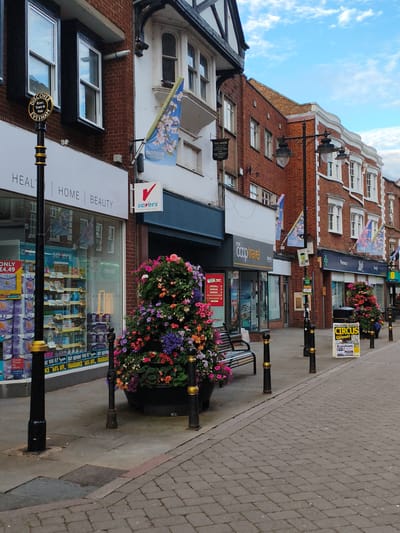 A street scene in Evesham, Wychavon, United Kingdom, features a row of shops with multi-story brick buildings. On the left, a black lamppost with gold accents stands in front of a store window displaying various products, including items with "ONLY £4.49" advertised. A sign above reads "HEALTH | HOME | BEAUTY." Further down the street, another lamppost is visible near a large planter filled with colorful flowers, including pink, purple, and orange blooms. Adjacent to the planter is a bench.
Moving right, a "Savers" store is indicated by a red and white sign. The next shop appears to be "Your Coop Travel." Further along, a "Pharmacy" sign is visible. Multiple flags with floral designs, possibly cherry blossoms, are displayed on poles along the street. A black trash bin is situated next to a sign advertising "CIRCUS Ginnett Evesham." The pavement is paved with gray bricks laid in a herringbone pattern. The sky above is blue with scattered white clouds. No people are visible in the frame. The scene appears to be an outdoor commercial area.