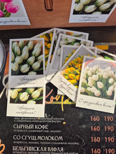 The image shows a collection of small cards with floral designs, predominantly white tulips, scattered on a table. The cards have messages in Russian, including "С праздником весны!" (Happy Spring Holiday!), "Пусть в душе всегда будет весна!" (Let there always be spring in your soul!), and "Цветущего настроения!" (Blooming mood!). Next to the cards is a menu with descriptions and prices of coffee drinks, including "Сырный кофе" (Cheese Coffee) and "Со сгущ молоком" (With condensed milk), as well as "Бельгийская вафля" (Belgian waffle). The prices are listed as 160 and 190, presumably rubles. The items suggest the location is likely a café or similar establishment in Neftekamsk, Russia, based on the Cyrillic text. The scene depicts a promotional display for spring or a set of greeting cards, placed in a café environment.