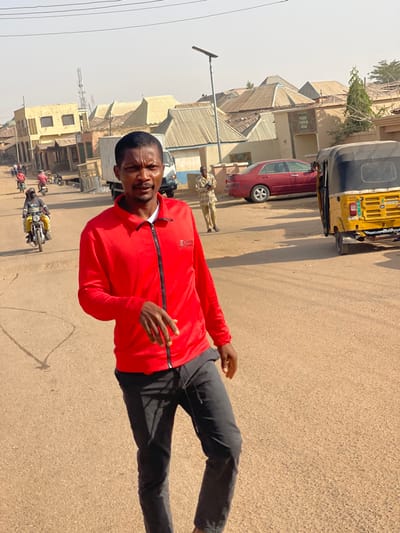 The scene depicts a daytime street view in Unguwar Gabas, Nigeria, under clear, hazy skies with strong sunlight casting distinct shadows.
In the foreground, a male subject, approximately 25-35 years old, stands facing the camera. He wears a red long-sleeved zipped track jacket with "SPORTS" text and a logo on the left chest area, and dark-colored trousers. His right hand is slightly extended downwards and open. He has short dark hair, a mustache, and a beard.
The midground features a dusty, unpaved road. To the right of the main subject, a yellow three-wheeled auto-rickshaw (keke napep) is partially visible, facing away, with a license plate partially obscured but showing elements like "JG114NG". Further back on the right, a red sedan with visible damage or dirt on its rear bumper is parked. A light-colored building behind the red sedan features "FADA SARKIN YAKI" written on its facade. A man in light-colored attire stands near the red sedan, appearing to hold a mobile phone. A solar-powered street light pole is visible between the auto-rickshaw and the red sedan.
To the left of the main subject and further down the road, two individuals on motorcycles are observed, one in yellow clothing on a yellow motorcycle, and another behind them. Multiple other pedestrians are faintly visible in the distance. A white box truck is partially visible between the main subject and the distant pedestrians.
The background consists of numerous one and two-story buildings with corrugated metal roofs, indicating residential and commercial structures common in the area. A tall communication tower or mast is visible in the far-left background.