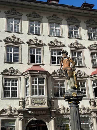 This image shows a multi-story building facade in Schaffhausen, Switzerland. The building is pale in color, possibly light gray or off-white, and features numerous rectangular windows with a grid-like pattern of panes. Each window is surrounded by ornate stone or plaster decorations. A statue of a man, possibly a soldier or historical figure, stands prominently in front of the building, mounted atop a column. The column features decorative elements like a golden flower and inscribed text that reads "BVRT CRISTI." There is also signage on the bottom of the building indicating a commercial space, likely including a hair salon or café. The street number "28" is visible near an arched opening. The sky is blue in the upper left corner.