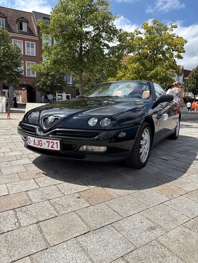The image is a low-angle outdoor shot of a black convertible Alfa Romeo Spider parked on a cobblestone plaza. The car is the primary subject, with its front grille, headlights, and license plate clearly visible. The license plate reads "0-AJG-721". Behind the car, there are buildings with red brick facades and pitched roofs, characteristic of European architecture. Trees with green and yellow foliage are also present, suggesting it is autumn or late summer. In the background, several people are visible, some walking and others standing, suggesting a public area or event. The sky is blue with scattered clouds. The scene appears to be a sunny day. The architecture, particularly the brickwork and window styles, along with the general ambiance, strongly suggests the historic city of Bruges, Belgium.