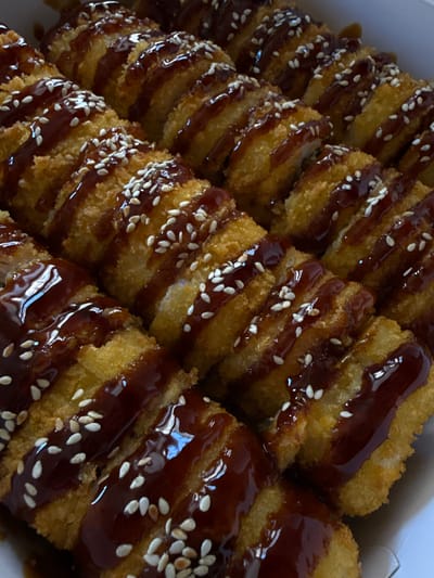 A close-up, overhead view shows a white cardboard container filled with several rows of golden-brown, fried sushi rolls. The rolls are cut into bite-sized pieces and arranged closely together. Each piece is drizzled with a dark, glossy sauce, possibly teriyaki or a similar glaze, and sprinkled with white sesame seeds. The lighting highlights the crispy texture of the sushi and the sheen of the sauce. The image does not contain any people, actions, or interactions. There is no visible information within the image to suggest its location in Hrazdan, Armenia, or any other specific city.