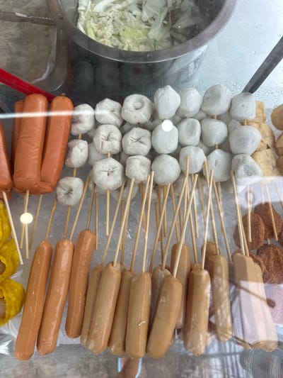 The image displays a close-up, overhead view of a food stall in Phuket City Municipality, Thailand. The stall appears to be a street food vendor, with various items displayed on skewers and in a metal container.

The primary subjects are food items arranged on wooden skewers, including white fish balls, pale brown sausages, and some fried brown items, possibly patties or more fish balls, at the top. In a circular metal container to the left, there is a pile of shredded white cabbage, likely for serving with the food. The food is laid out on a bed of ice, suggesting it is being kept cold.

The scene is brightly lit, likely indicating it is daytime, though no specific time can be determined. There are reflections on the glass or plastic covering the food, indicating the vendor is enclosed. The overall impression is of a vibrant and busy food stall, typical of street food scenes in Thailand. No people are visible in the frame. There is no visible text in the image. The activity taking place is the preparation and display of food for sale.