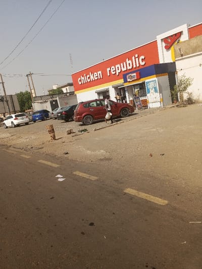 The image depicts an exterior scene in Gawuna, Nigeria, featuring a "Chicken Republic" fast-food restaurant. In the foreground, a dark asphalt road with faded yellow dashed lane markings contains scattered white paper and dark stains. To the right of the road, a dirt and gravel shoulder transitions to a parking area. A short, thick wooden post or stump stands on this shoulder.
In the midground, a dark red crossover SUV is parked facing left on the dirt lot. A dark-skinned individual wearing a white shirt and dark pants walks left, carrying a white bucket in their left hand, positioned next to the red SUV. Beyond this vehicle, several other cars are parked in the unpaved lot, including a white sedan, a blue sedan, and a dark-colored sedan.
The background is dominated by the single-story "Chicken Republic" building, which features a prominent red fascia with "chicken republic" in large white lettering. A yellow and white "Y" logo is visible on the upper right section of the building. The entrance area is recessed, with blue and yellow architectural accents. Several individuals are visible near the glass entrance doors, and a promotional sign for "SHAWARMA & SHAWARMA SNACKS" is displayed to the right of the entrance. A small green shrub grows near the building's right side. Utility poles with overhead lines are present on the left side of the scene, and a communication tower is visible in the distant background under a clear, light blue sky. The overall environment appears dry and dusty.