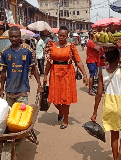 A woman in an orange dress with a black belt walks through a busy market. She carries a black purse. A young man pulls a wheelbarrow with a yellow container and a white bag. Another person carries bananas on their head. People and stalls fill the background, shaded by umbrellas. Buildings are visible in the distance. The ground appears dusty.