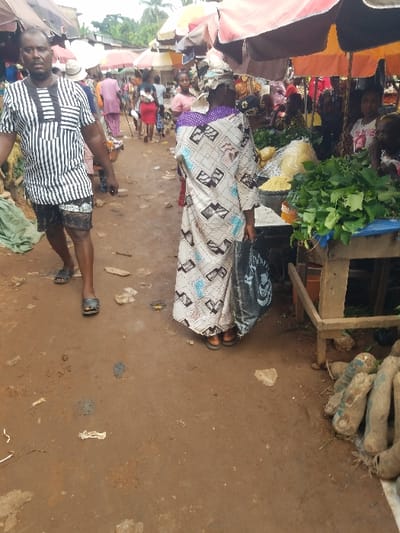 This image captures a bustling outdoor market scene in Nnobi, Nigeria. The foreground features a reddish-brown dirt path scattered with minor debris.
On the left, a dark-skinned male subject walks towards the viewer, slightly off-center. He has short dark hair and a mustache, wearing a short-sleeved, collared shirt with alternating black and white vertical stripes and a geometric pattern on the chest. He wears patterned dark shorts and dark open-toed sandals.
In the center, a female subject is viewed from behind, walking away from the camera. She wears a patterned headwrap with purple accents and a loose-fitting, long-sleeved dress or gown in a light color with a repeating geometric pattern in black, white, and light blue. She carries a dark shopping bag in her left hand and wears dark open-toed sandals.
To the right, a rustic wooden market stall displays produce. A large pile of fresh green leafy vegetables rests on a blue cloth. To their left, a pile of yellow granular substance, possibly garri, is visible in a clear plastic bag. On the ground next to the stall, several elongated, light brown tubers, likely yams, are arranged, partially covered by a white cloth.
The midground and background show numerous other market stalls with colorful large umbrellas providing shade, predominantly in red, orange, brown, and beige. Many people are visible moving through the market, indicating active commerce. Green foliage and tree tops are discernible above the market structures in the distant background. The overall lighting is soft and even, consistent with an overcast day.