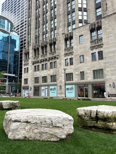 This is a vertical, eye-level, full shot of a grassy area with large rocks in the foreground, with a classic stone building and a modern glass building in the background. The grassy area is well-maintained and appears to be part of a park or urban plaza. Two large, light-colored boulders are the most prominent objects in the foreground, with a third smaller boulder visible to the left. The larger boulder on the right has a rough texture and appears weathered, with some green moss or lichen on its surface.
Behind the rocks, a tall, ornate stone building stands prominently. Its facade is characterized by a repetitive pattern of windows and intricate architectural details, suggesting it is an older, possibly historic, structure. Several signs are visible on this building, including two identical signs that read "BIG CITY OPTICAL Coming Soon".
To the left of the stone building, a modern glass skyscraper rises, its reflective surface showing the sky and surrounding buildings. The contrast between the old and new architecture is striking.
Several people are visible in the scene, mostly in the background near the buildings, suggesting a daytime setting. Some appear to be walking, while others are engaged in activities that are not clearly discernible due to the distance and image orientation. The weather appears to be overcast or the light is diffused, as there are no harsh shadows. The overall impression is an urban environment where nature, represented by the grass and rocks, has been integrated with architecture. The presence of the "Coming Soon" signs hints at a developing commercial area.