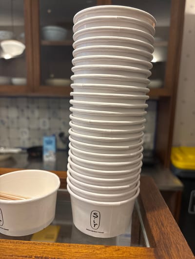 A stack of white paper cups with ridged rims sits on a glass surface in front of a wooden counter. The cups are a branded product, with the logo "S'r" and the text "GREEK FROZEN YOGURT" visible on the bottom cup. To the left of the stack, a single white paper bowl containing two wooden stirrers is visible. In the background, a wooden cabinet with glass doors displays various bowls and a blue carton. The setting appears to be a food establishment in South Jakarta, Indonesia, possibly a cafe or yogurt shop.