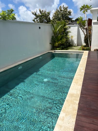 The image captures a swimming pool in Ungasan, Indonesia. The pool's water appears turquoise and ripples with sunlight. A wooden deck borders one side of the pool, separated by a light-colored stone border. On the opposite side, a white wall runs alongside the pool, partially obscured by tropical foliage, including palm trees. The wall is adorned with a small, square dark fixture. A glimpse of the sky reveals blue patches and fluffy white clouds. Other buildings and vegetation appear in the background.