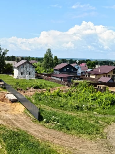 The image depicts a suburban landscape in Yaransk, Russia, on a clear, sunny day with scattered white clouds. The foreground features a dirt path with tire tracks and a pile of concrete blocks next to a dark metal fence. Beyond the fence, a newly constructed house made of light-colored cinder blocks is visible, surrounded by a green lawn. Several other houses with dark brown roofs are scattered throughout the midground, separated by wooden fences and lush green vegetation. The background consists of a dense line of trees, with some buildings and a distant church steeple peeking through. The overall scene suggests a developing residential area.