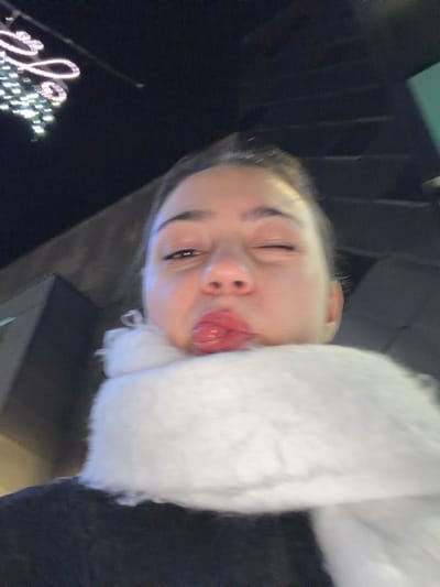 A woman angles her face, tilting her head and winking one eye at the camera. She is wearing a fluffy, white scarf. Her tongue sticks out playfully. The background consists of dark structures and a blurred string of lights, possibly holiday decorations. The lighting in the image appears low-light and the camera is positioned at a low angle, looking upwards at the subject.