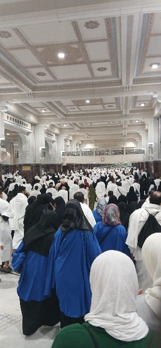 The image captures a dense crowd of individuals, primarily pilgrims, within a grand, brightly illuminated indoor hall located in Makkah Al Mukarramah, Saudi Arabia. The hall features an elaborate, multi-tiered coffered ceiling with decorative beige and white patterns and integrated square and circular recessed lighting fixtures. Tall white classical columns and arches line the spacious interior, while mid-height walls exhibit dark lower panels topped by intricate, light-colored balustrades or grilles with geometric designs. The floor is covered with light-toned tiles or marble.
The multitude of people, numbering in the hundreds and extending deep into the background, appear to be moving or standing in an orderly fashion. Numerous men are dressed in the distinctive white ihram garments, signifying their pilgrim status. Women are seen wearing various forms of modest attire, including black abayas and head coverings (hijabs) in colors such as white, black, grey, blue, and olive green. In the immediate foreground, a woman is seen from behind, wearing a green top and a white hijab. Slightly ahead of her, two women are visible from the back, dressed in bright blue long-sleeved tunics worn over dark lower garments, with black head coverings. Some individuals carry small backpacks. No specific interactions between individuals are discernible due to the crowd density.