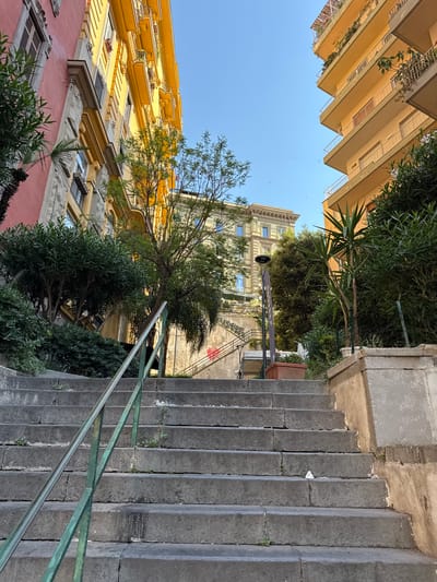 The image depicts a narrow, upward-sloping stone staircase in Naples, Italy. The staircase is lined with stone walls and flanked by buildings. To the left, a building with a pink facade and yellow upper stories is visible, with windows and balconies. To the right, a tall, light-yellow apartment building with multiple balconies and greenery growing on them dominates the view. Lush green foliage and trees are interspersed throughout the scene, with a palm tree to the right and smaller bushes and trees on the left. In the background, another building with a light-colored facade and arched windows can be seen, along with more greenery and a flight of stairs leading upwards. A dark lamppost stands near the center of the image. The sky is clear and blue. The scene appears to be a residential street or a pathway in a hilly part of the city.