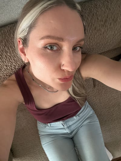 A young woman lies on a brown textured surface, possibly a couch or bed. She is facing upwards, with her head slightly tilted. She has fair skin, light blonde hair with darker roots, and green eyes. She is wearing a maroon tank top, a silver chain necklace, light blue jeans, and a silver nose ring. The background is blurred, but suggests a domestic setting. The location in Esenyurt, Turkey is not discernible from the image.